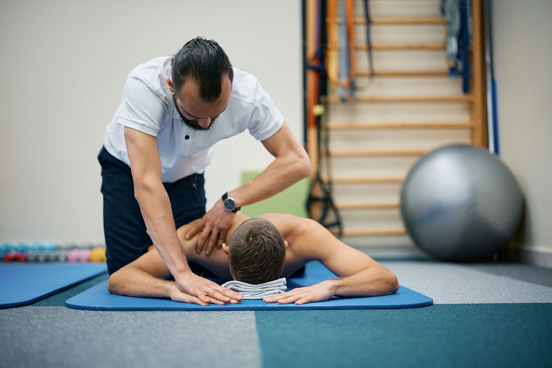 A therapist massages a person's upper back, who lies on a mat in a rehabilitation room.