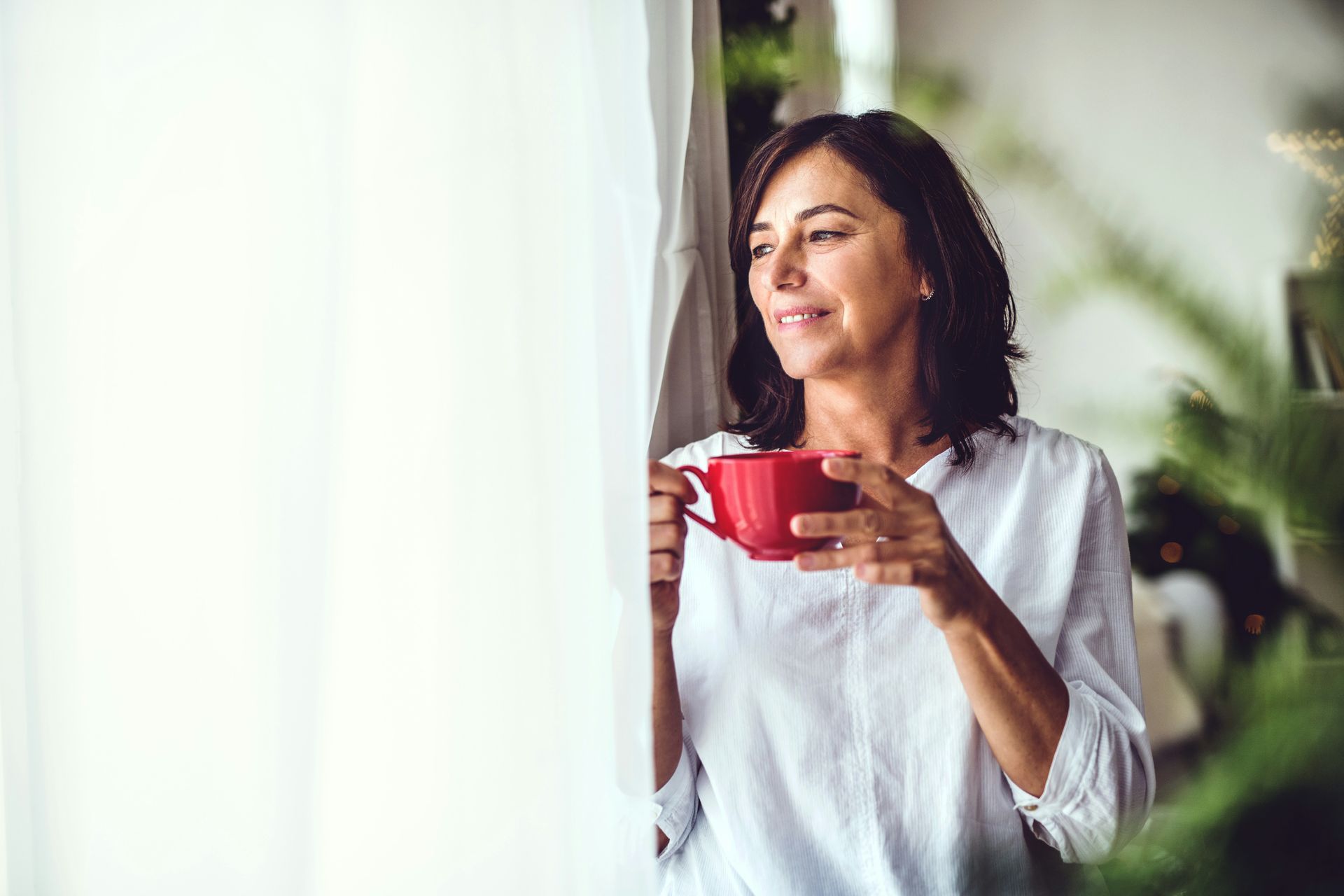 Woman with a red mug looks out window, holding white curtain.