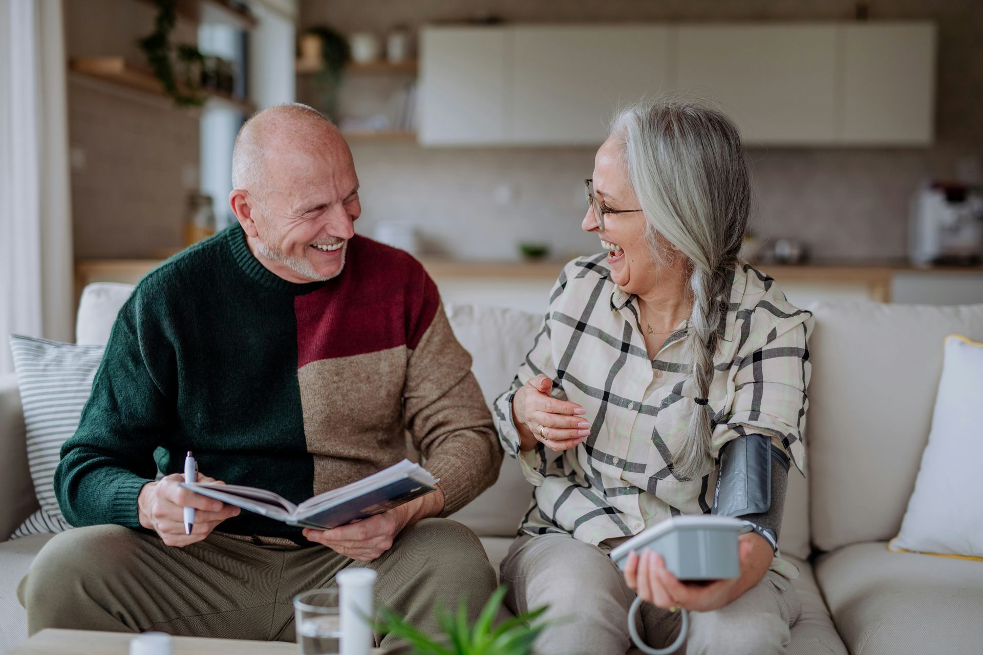 An older person sits on a couch, measuring their blood pressure while smiling.