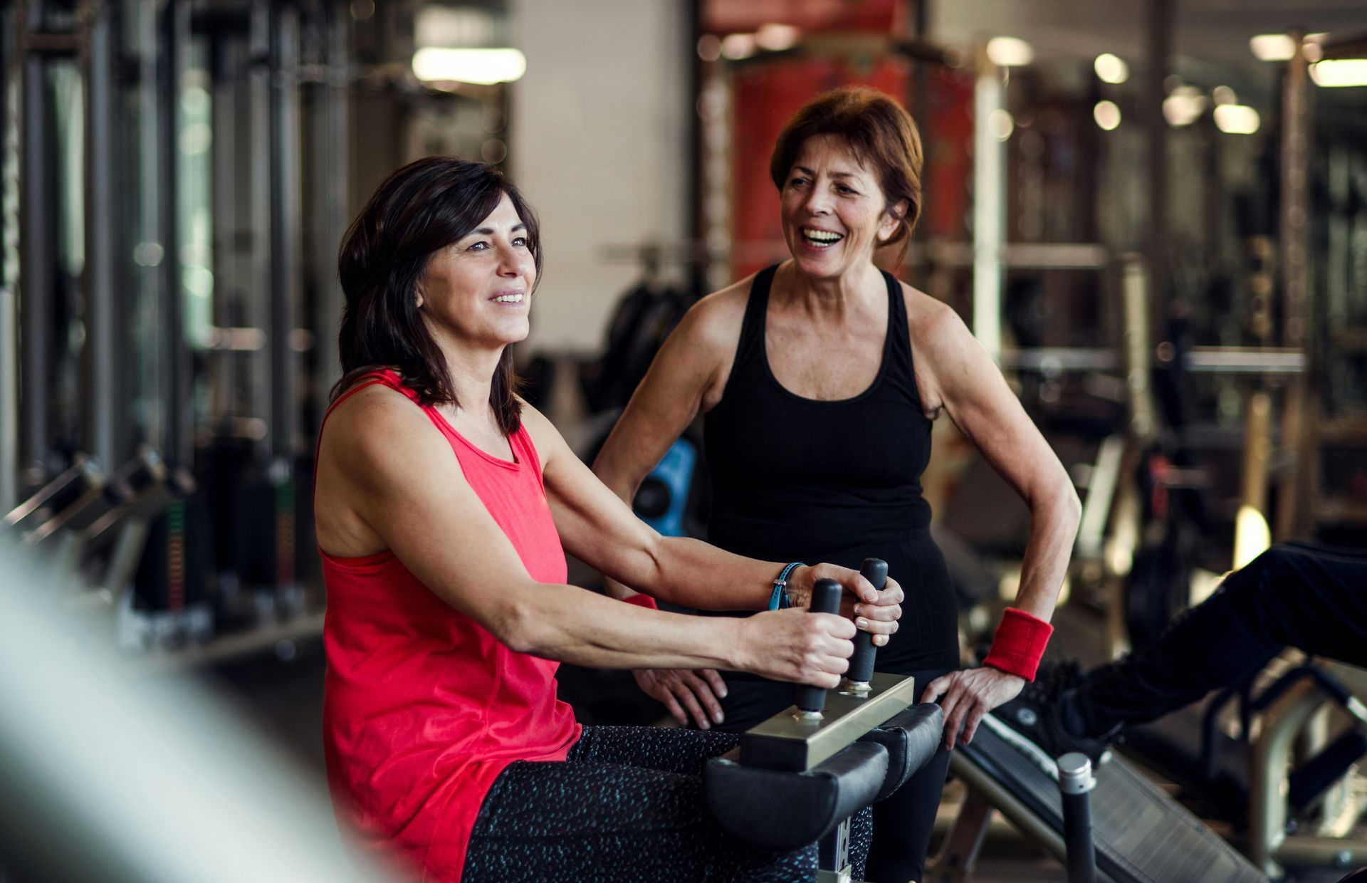 Two women at a gym; one uses a machine, the other smiles and watches. Red and black tops; gym setting.