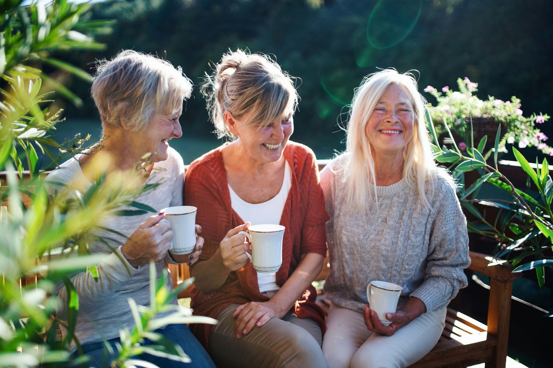Three women on a bench laughing, holding mugs, outdoors with greenery.