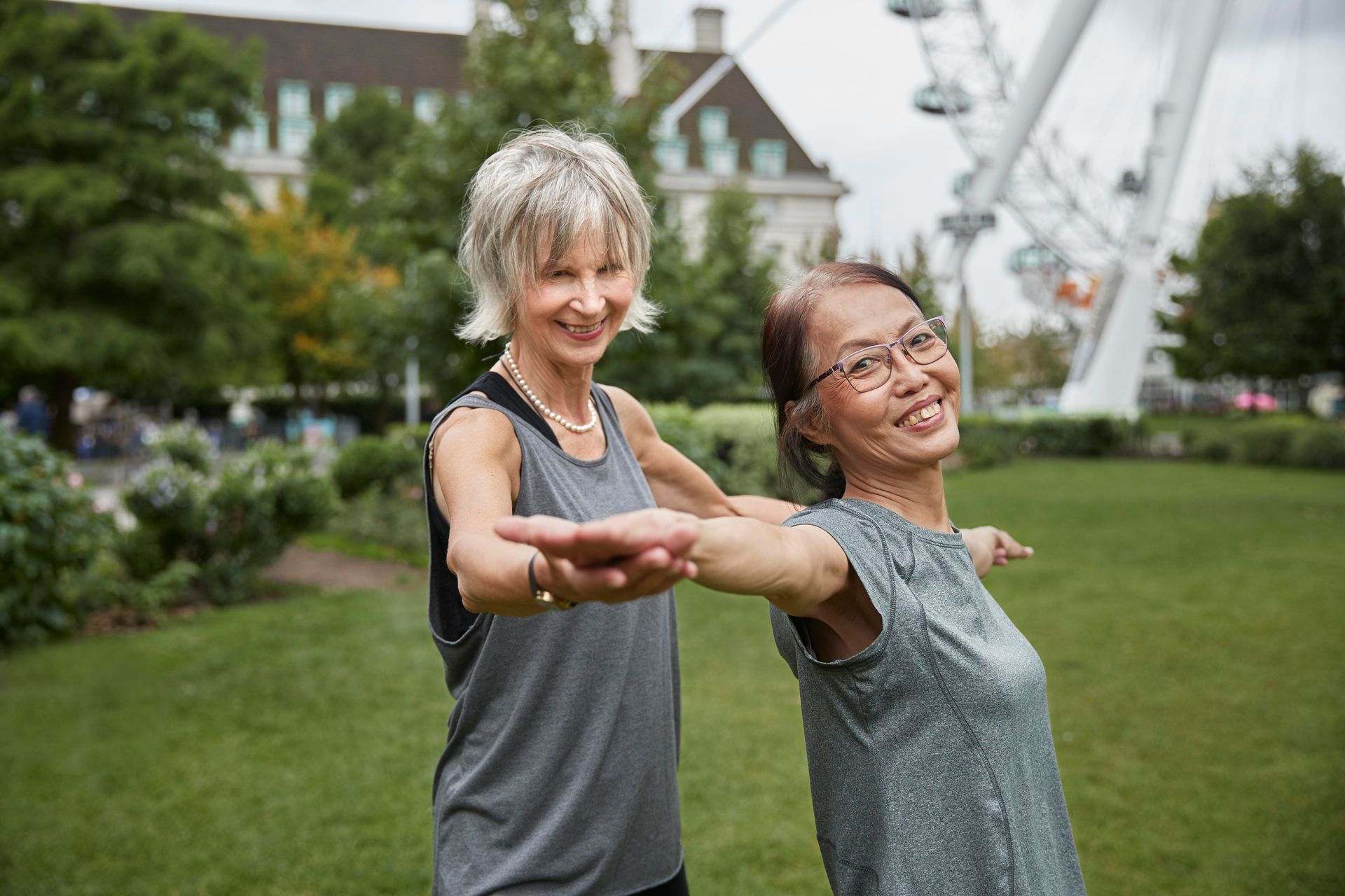 Two women stretching arms in a park, smiling. Ferris wheel in background.