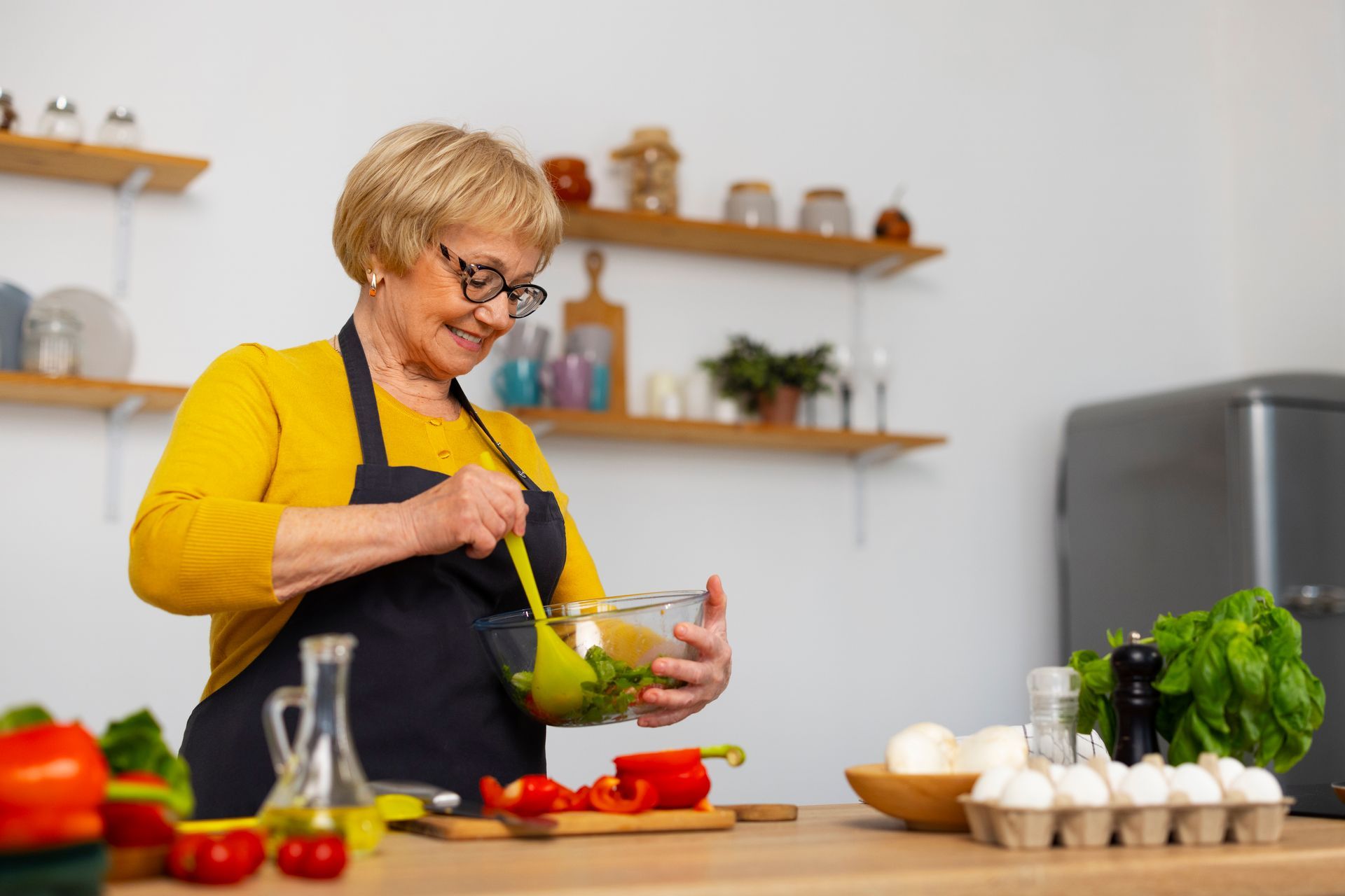 Woman in glasses wearing an apron stirring salad in kitchen. Red pepper and vegetables on counter.