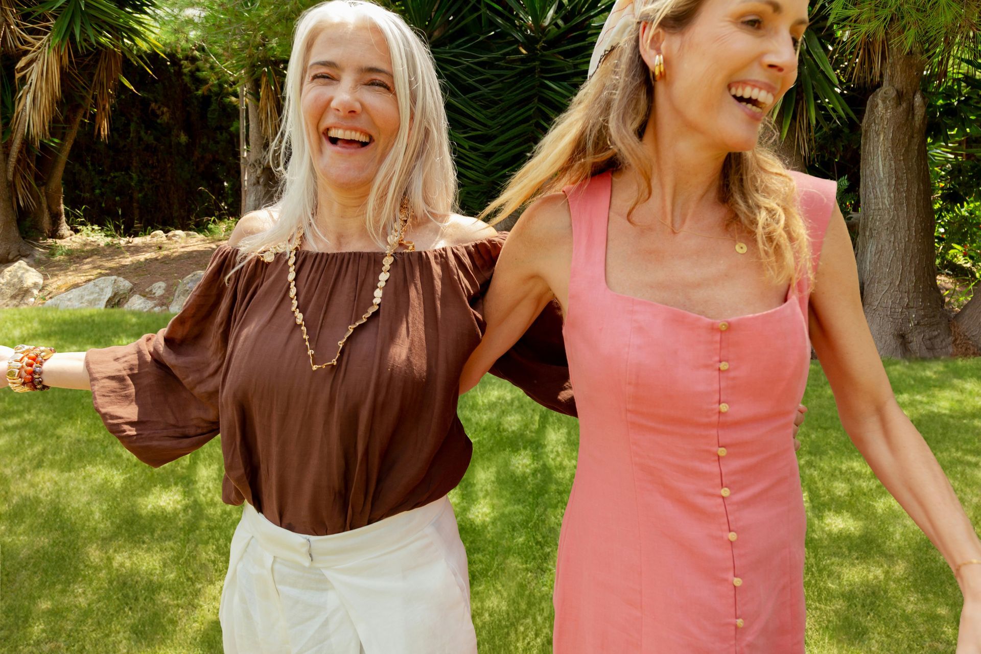 Two women laughing outdoors, arms around each other. One wears a brown top and white pants, the other a pink dress.