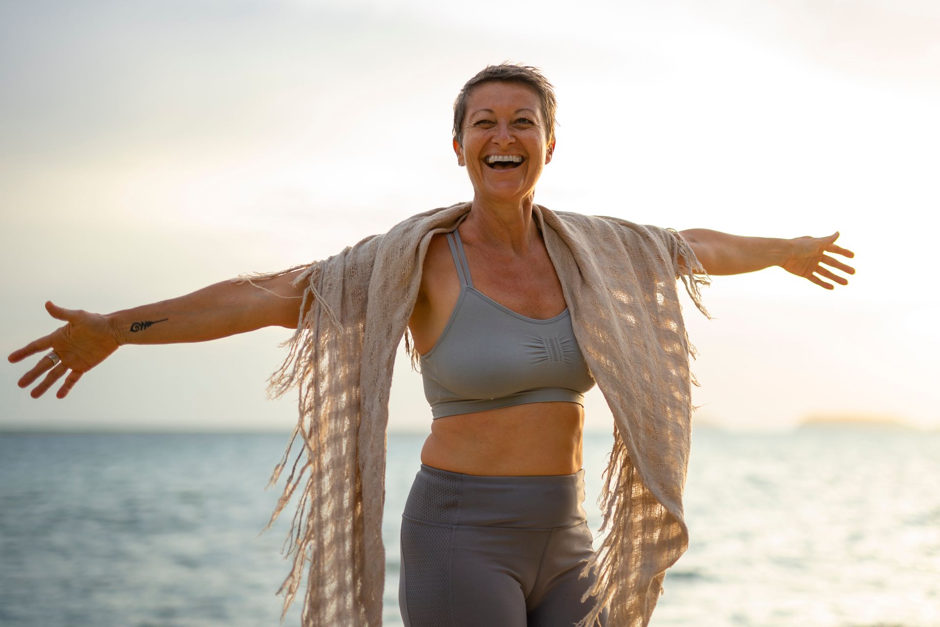 Woman with arms outstretched, smiling at the beach, wearing workout clothes and a shawl.