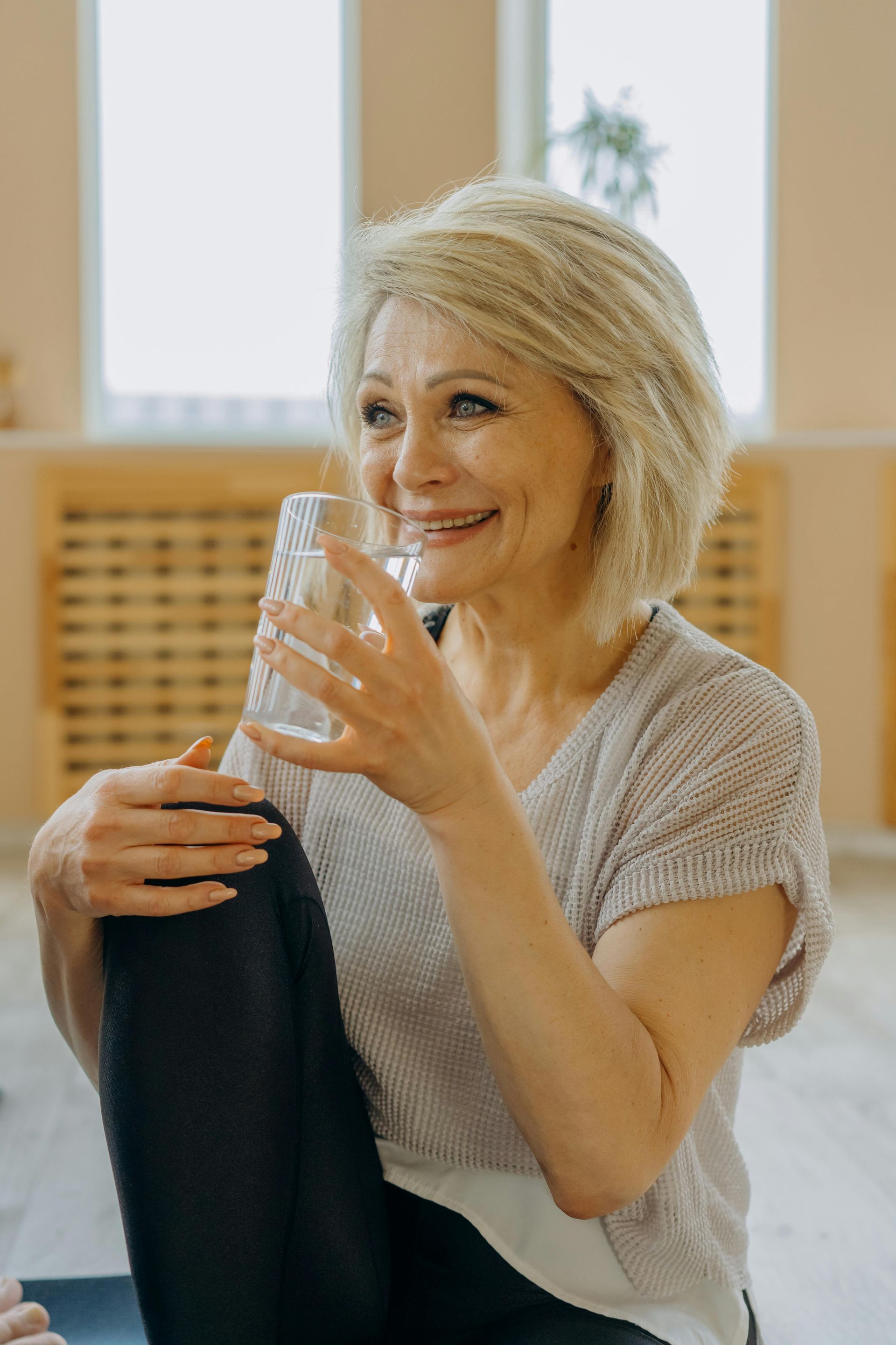 Woman with short gray hair smiles, holding glass of water, leg bent, sitting indoors.