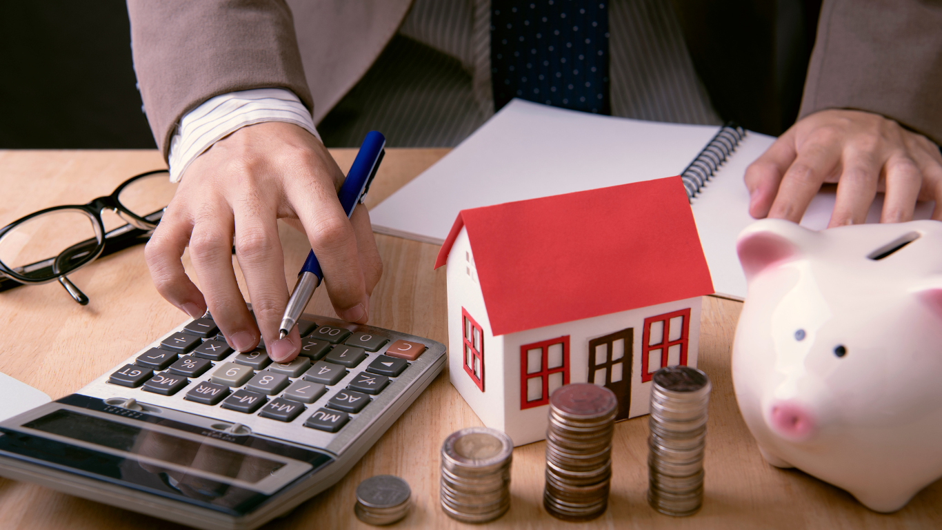 A man is using a calculator next to a piggy bank and a model house.