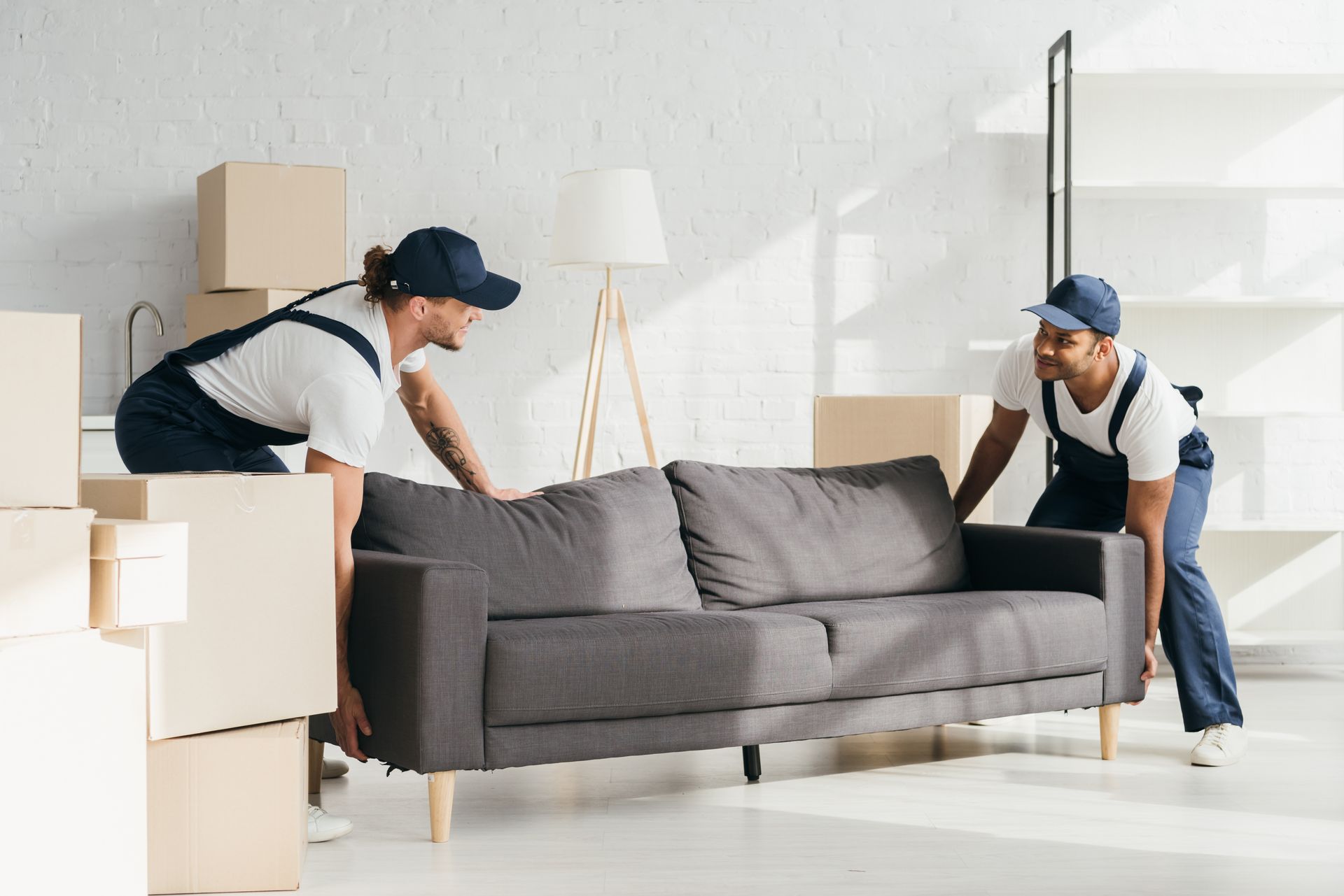 Professional movers are carefully lifting a heavy grey sofa in a brightly lit new apartment.