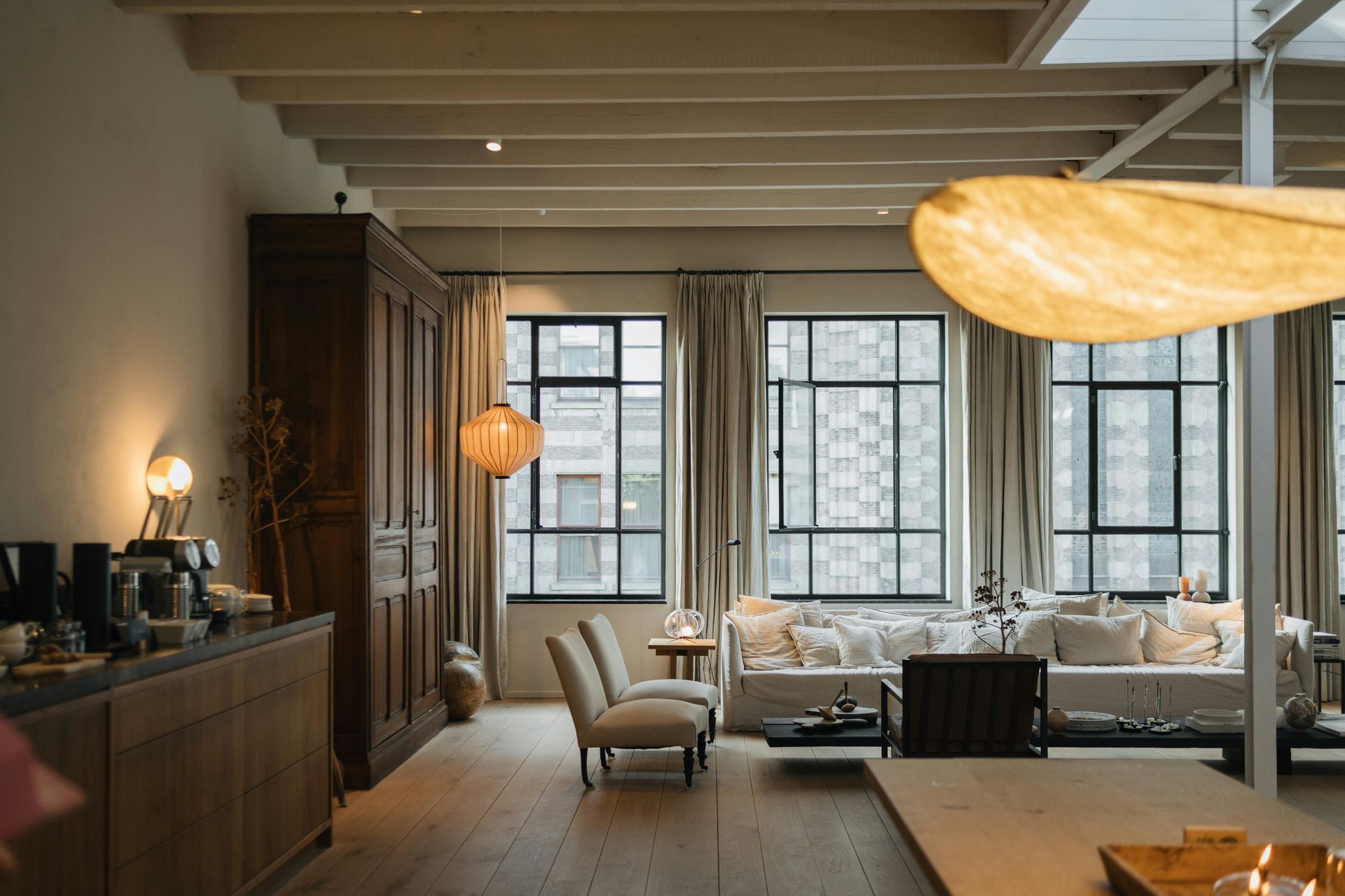 Spacious, neutral-toned living room with windows, seating area, wooden furniture, and decorative lighting.