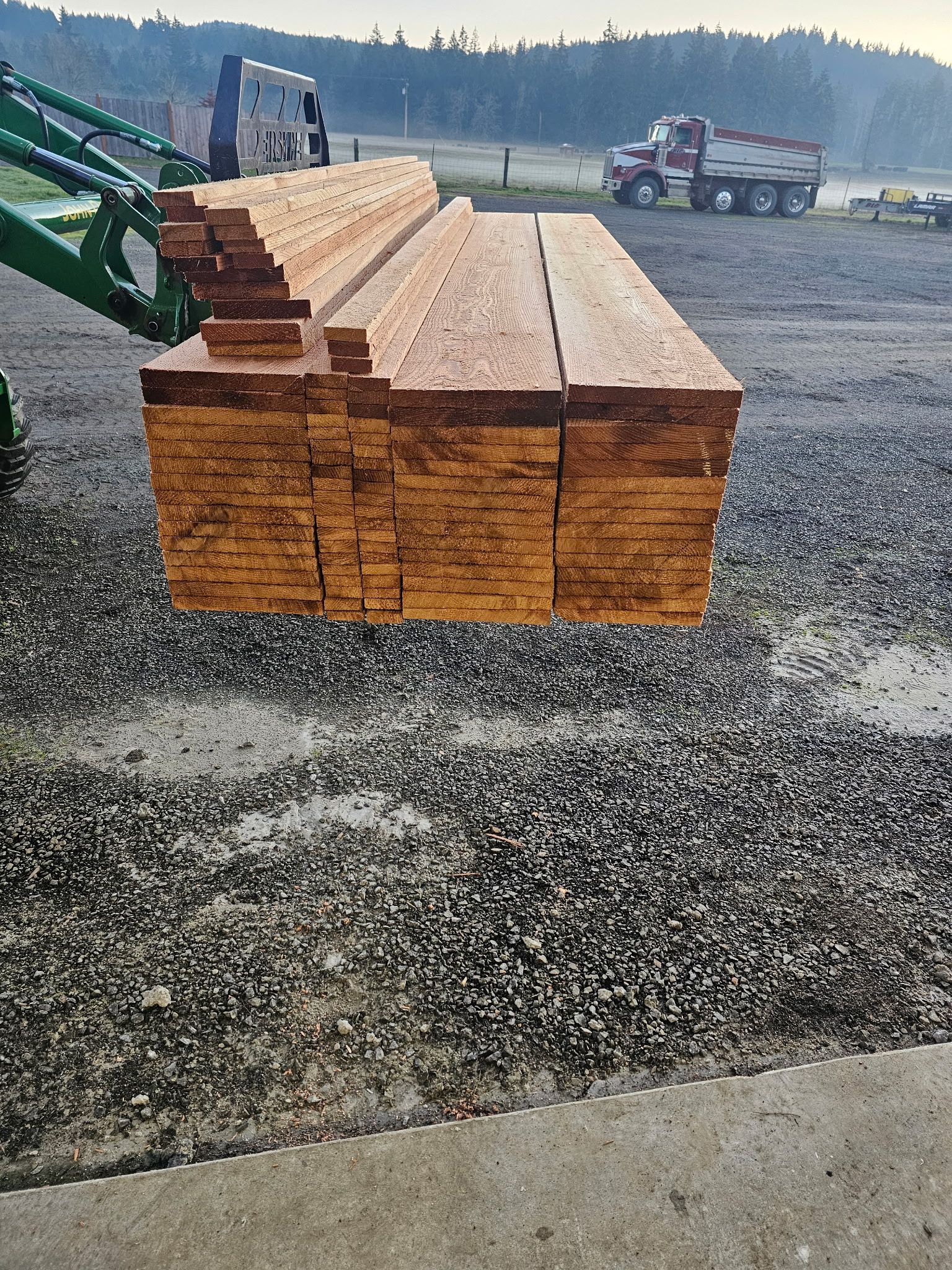 Forklift carrying a stack of lumber outdoors, with trucks and trees in the background.