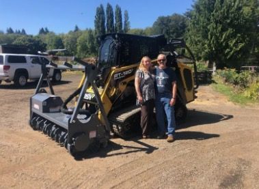 A couple stands with a yellow and black compact track loader with a brush cutter attachment. Sunny outdoor setting.