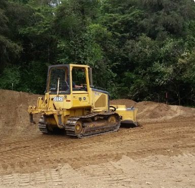 Yellow bulldozer smoothing dirt, in front of a treeline.
