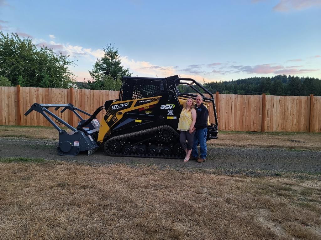 Couple stands with a yellow and black tracked skid steer with a forestry mulcher attachment. Outdoor setting.