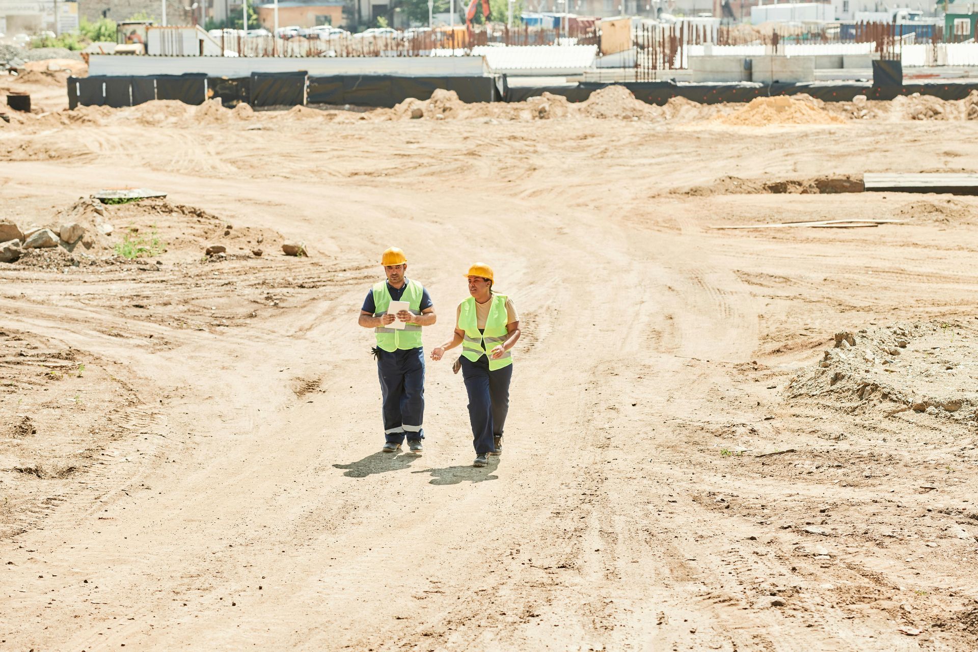 Two construction workers in safety vests walk on a dusty construction site.
