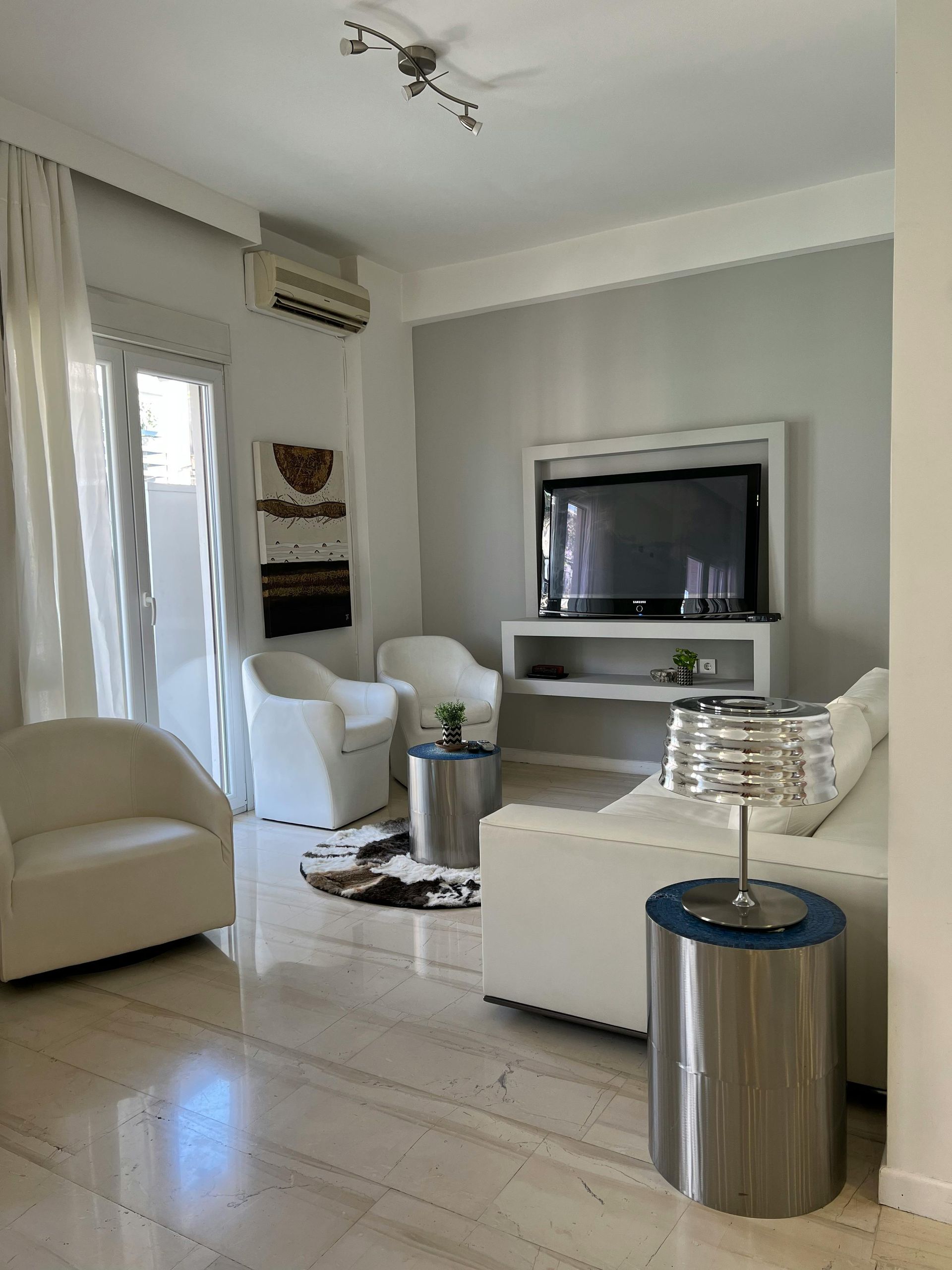 Bright, modern living room with white furniture, silver accents, and a large TV.