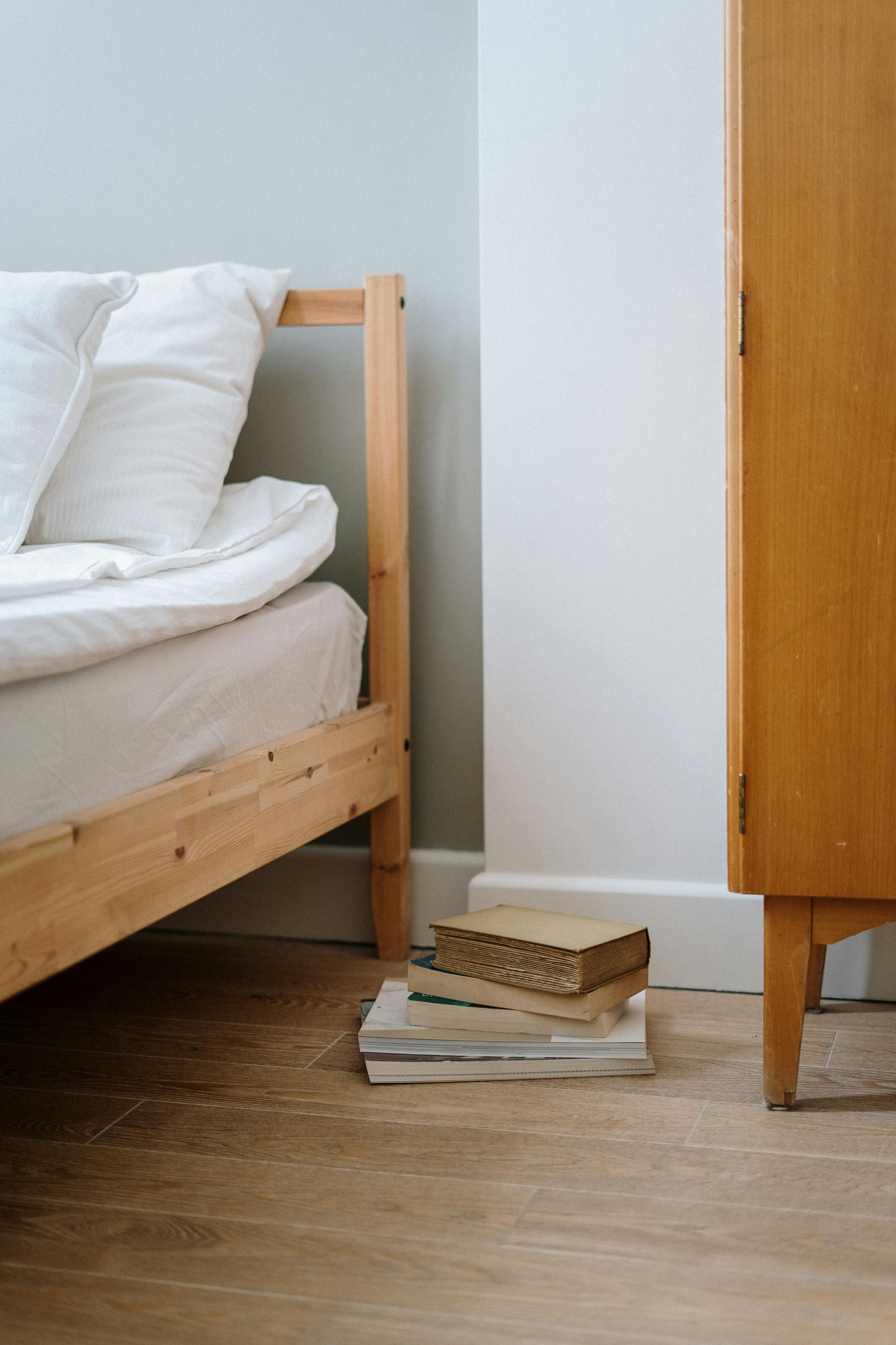 Wooden bed frame and wardrobe beside a stack of books and a box on a light wood floor.