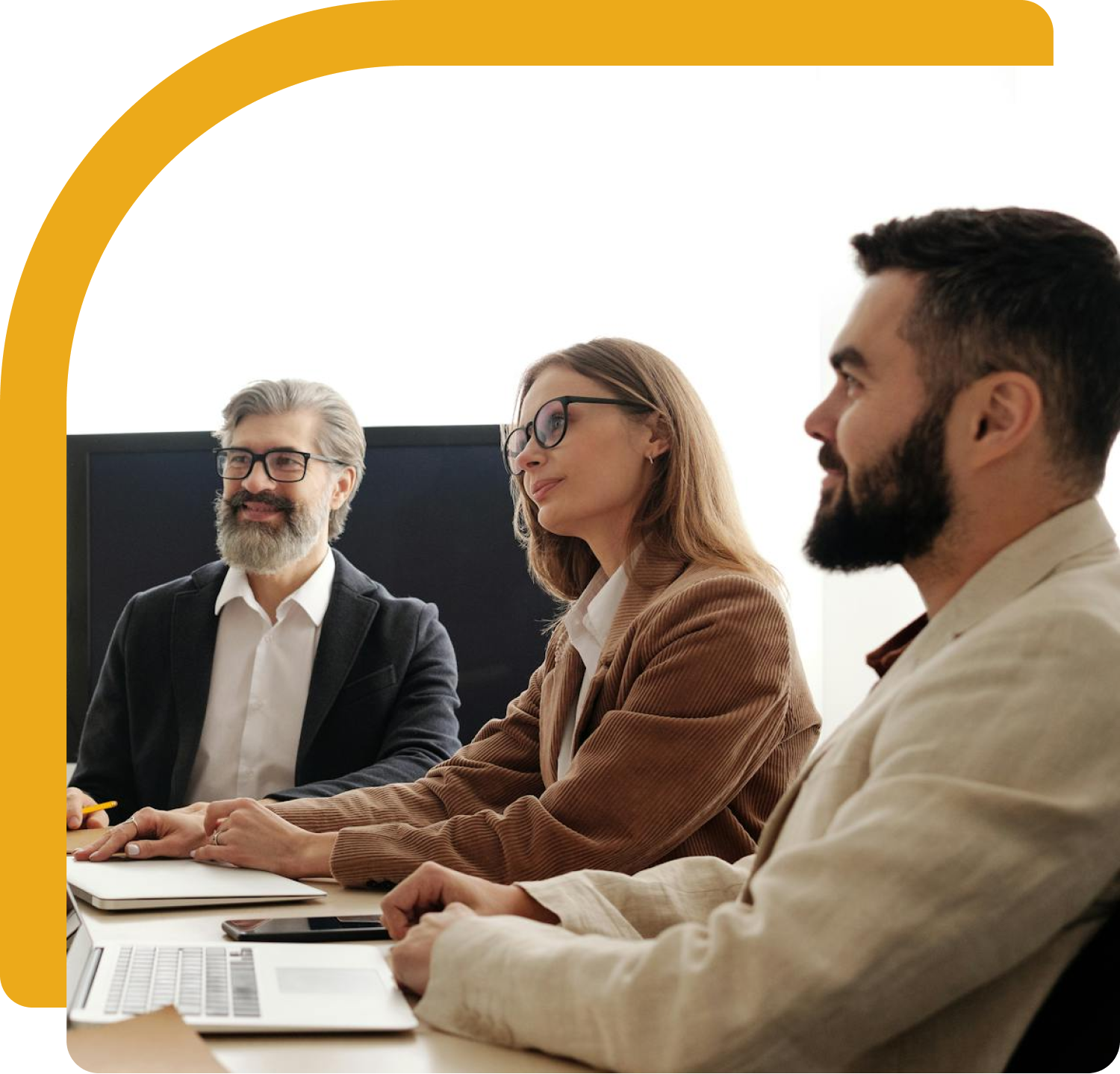 Three people in a meeting: a man with a beard smiles, and two others look at the table.