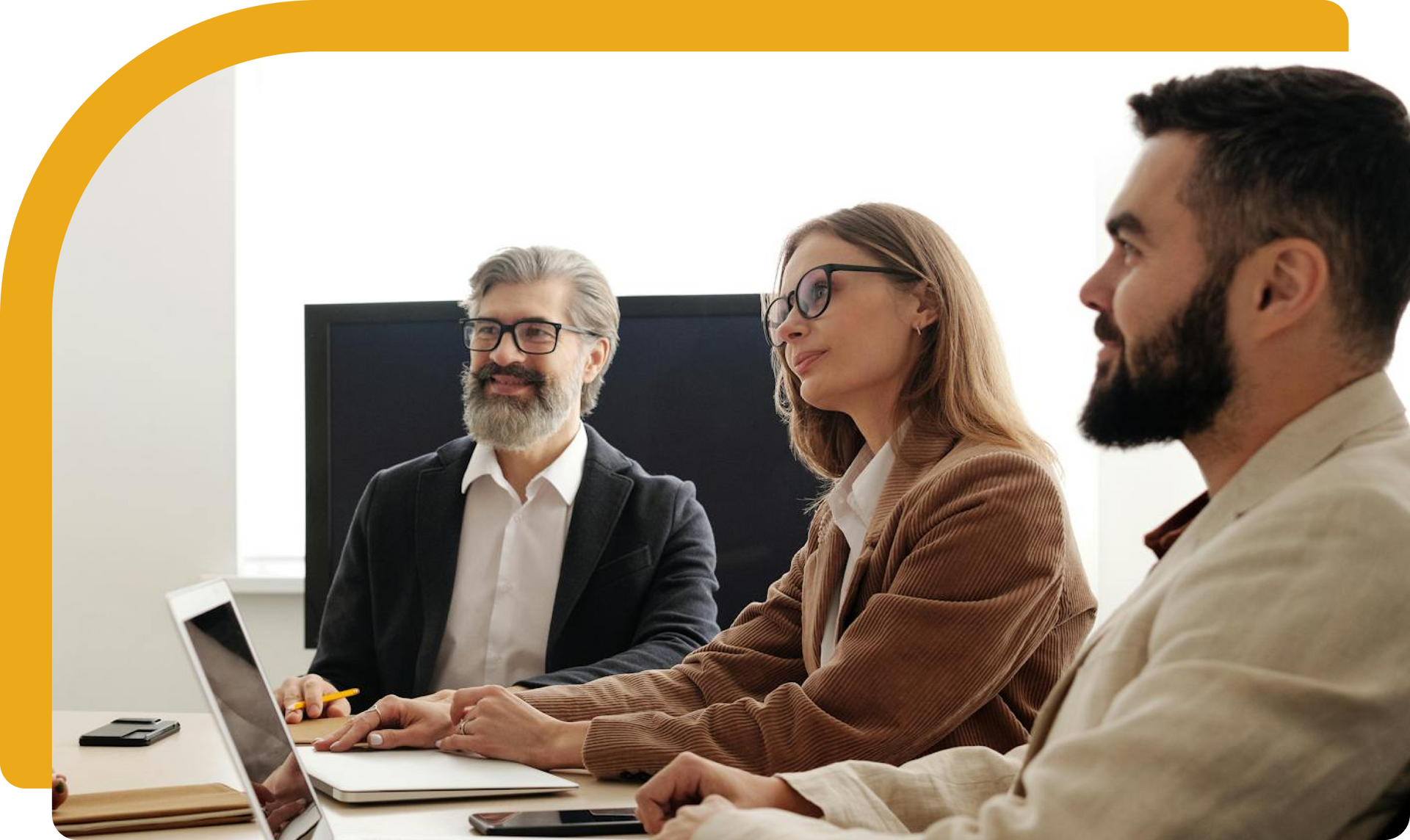 Three people in a meeting: a man with a beard smiles, and two others look at the table.