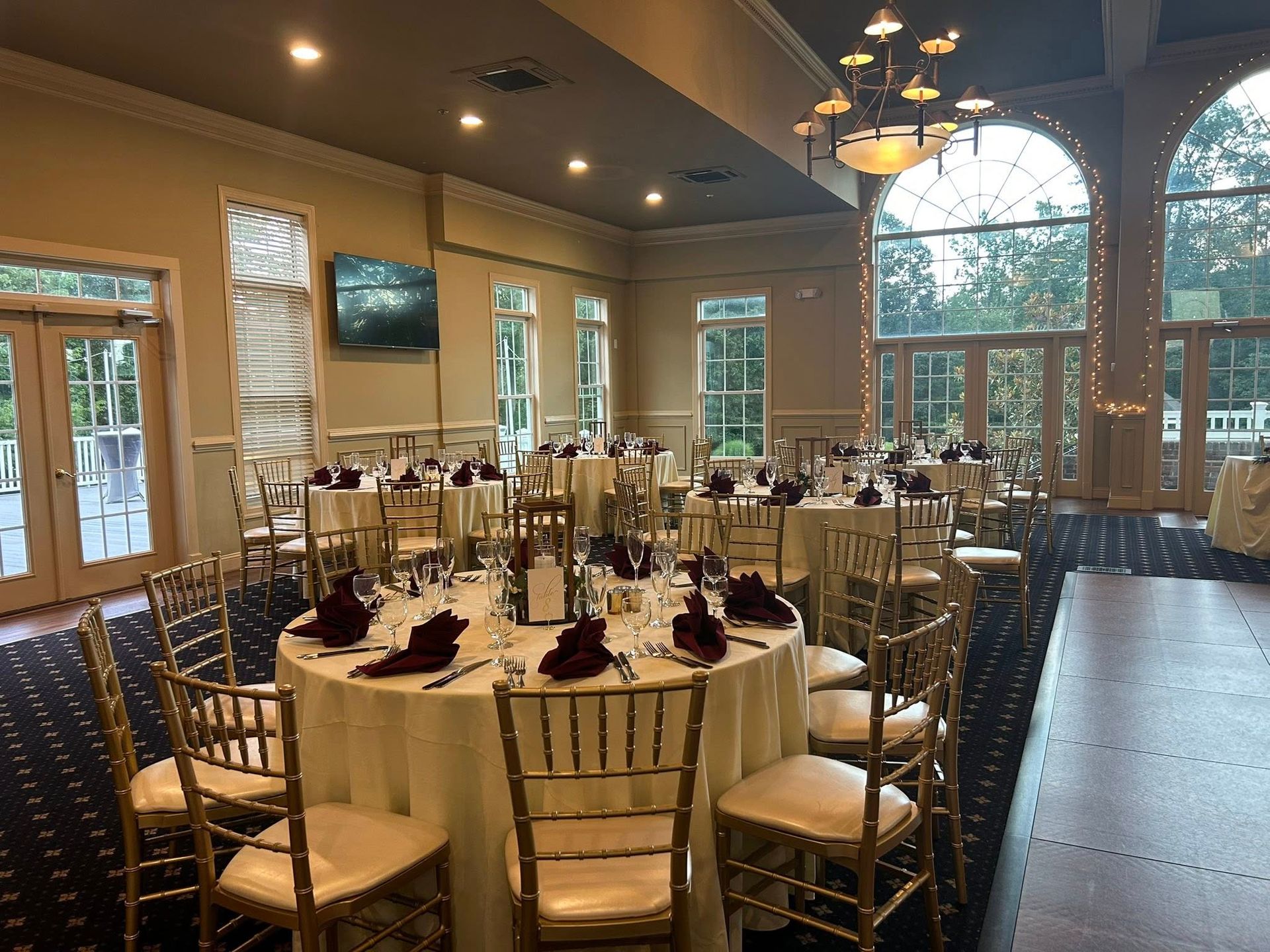 Wedding reception hall with round tables set for guests; gold chairs, burgundy napkins, and large windows.