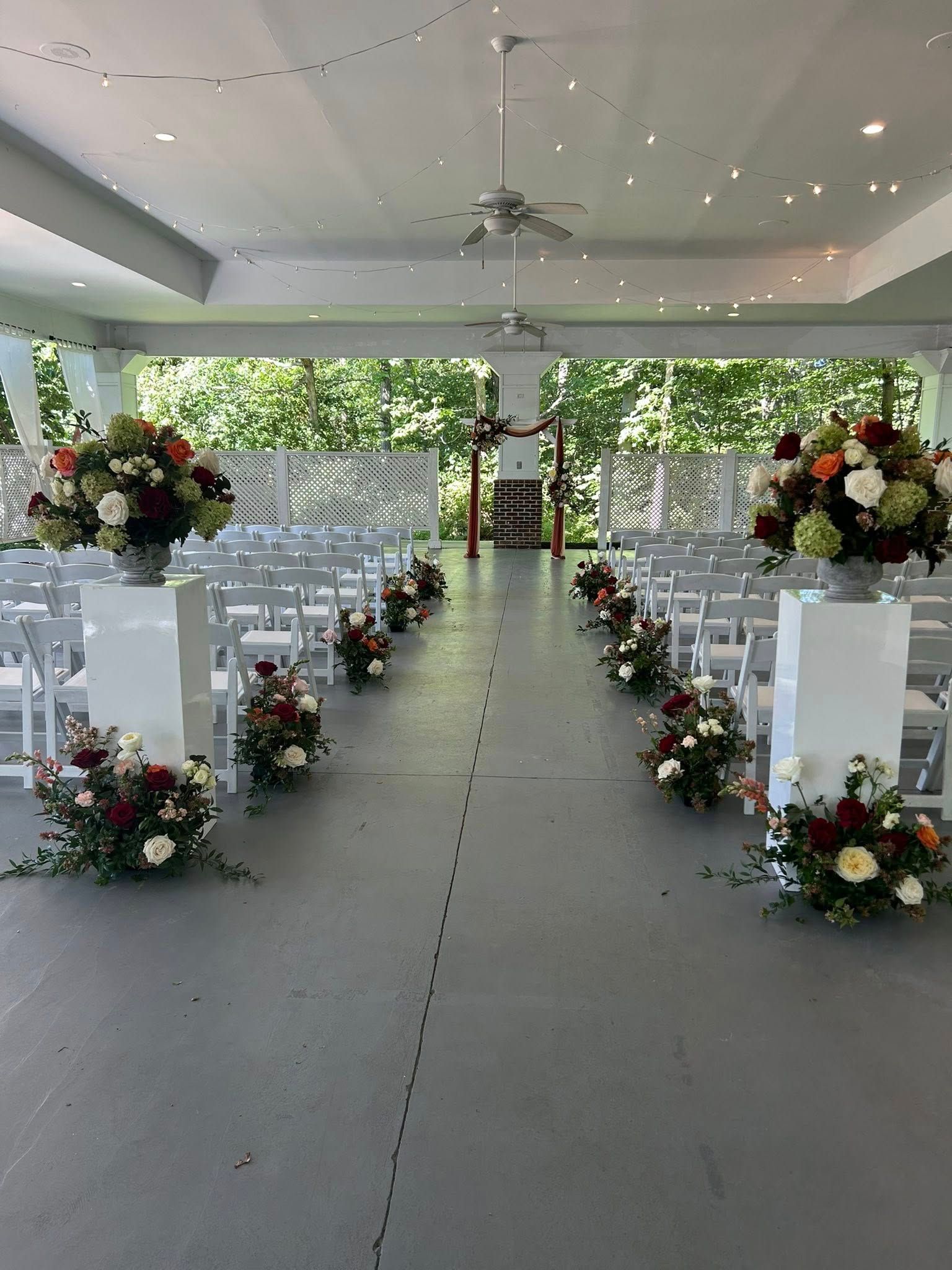 Wedding ceremony setup: White chairs line an aisle with floral arrangements. Green foliage outside windows.