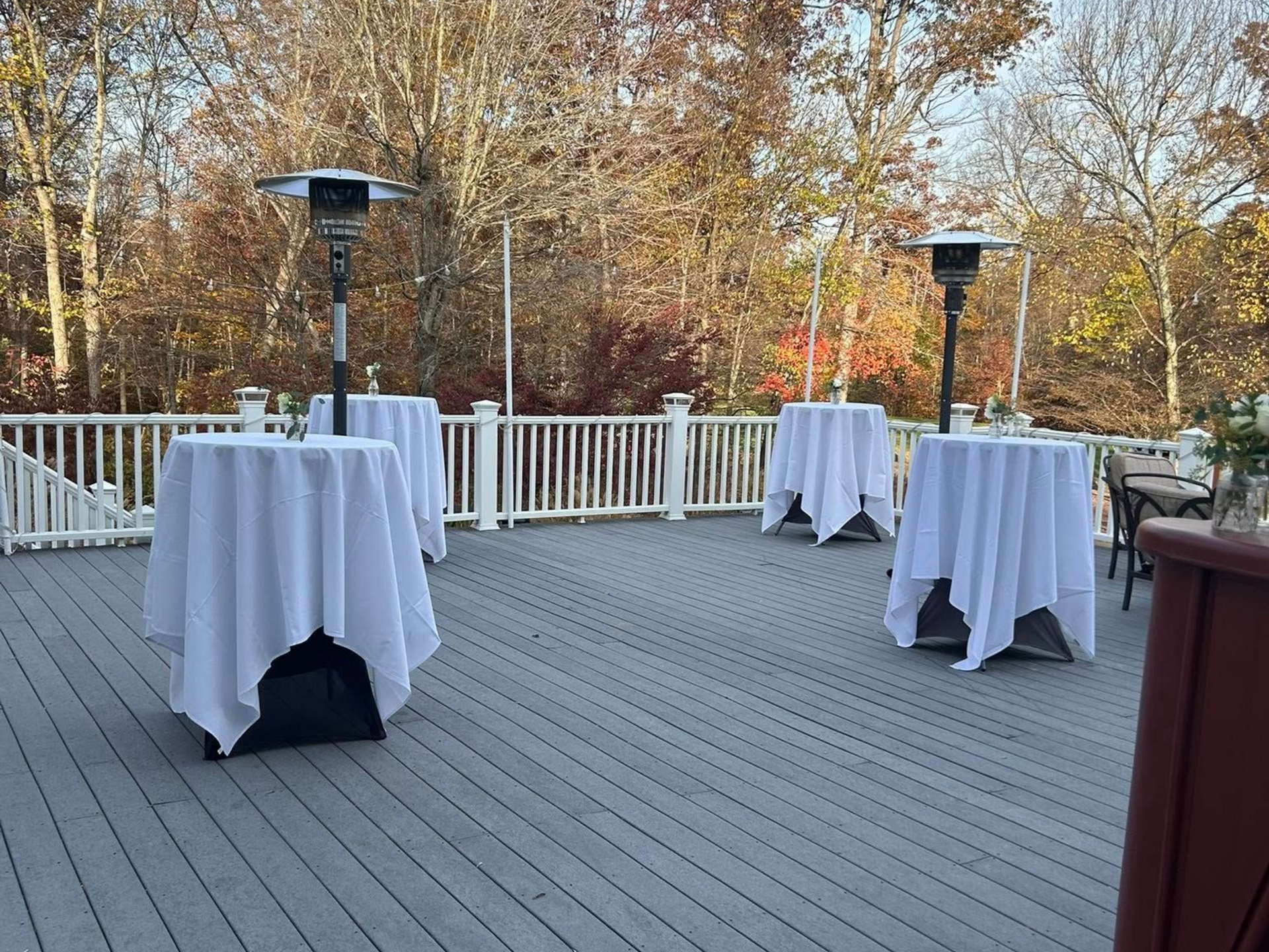 Outdoor deck with four white-covered tables, two patio heaters, and fall foliage in the background.