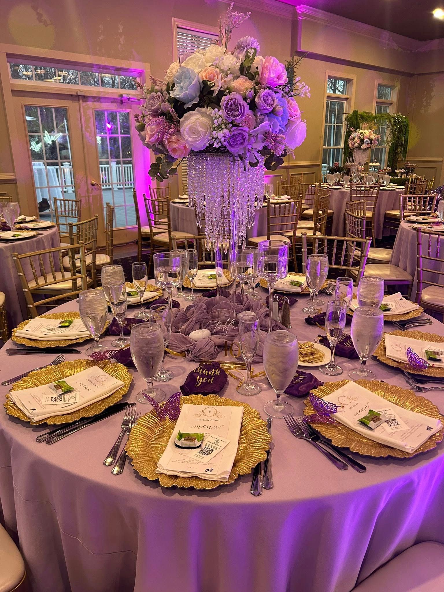 Elegant round table set for a formal event, featuring gold chargers, floral centerpiece, and purple lighting.