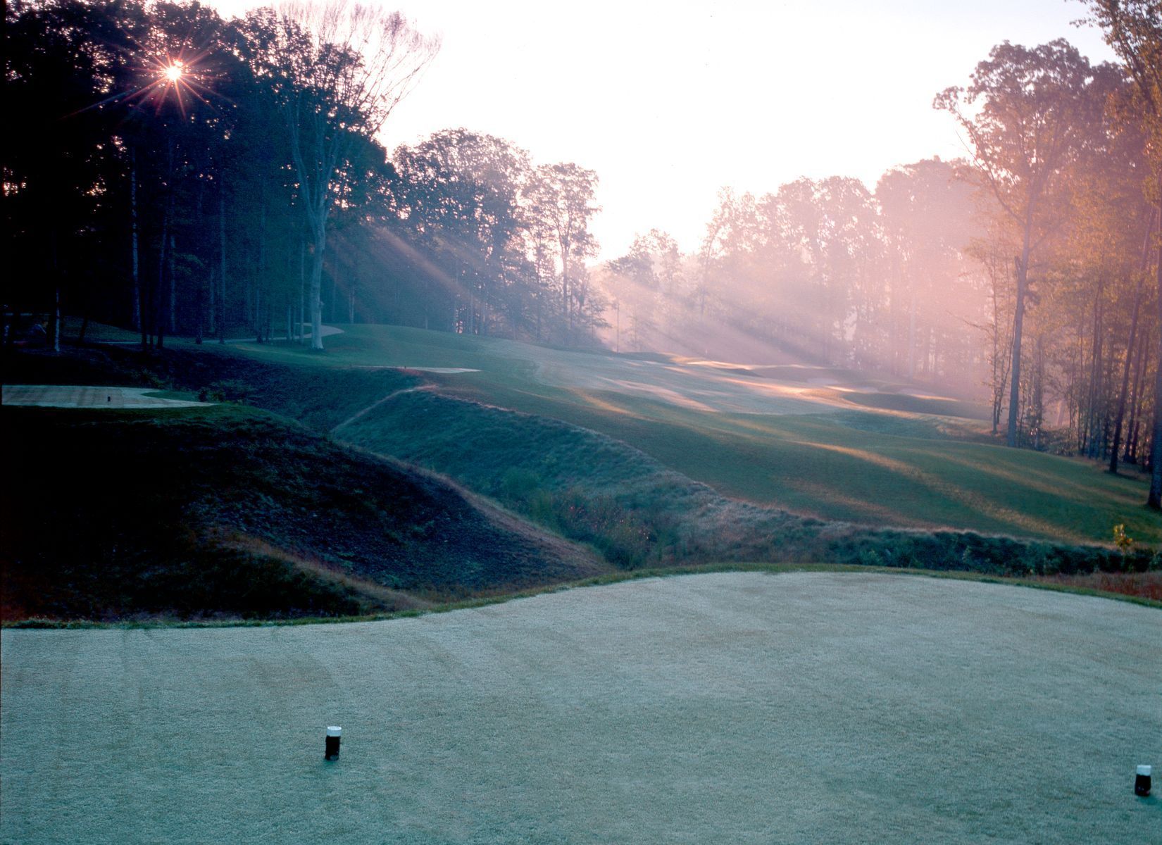 Golf course fairway at sunrise with sun rays streaming through trees.