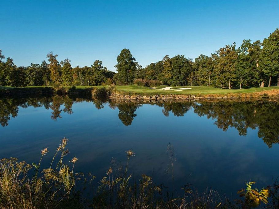 A calm pond reflects trees and a golf course under a clear blue sky.