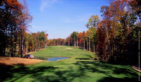 Green golf course with pond, surrounded by autumn trees under a blue sky.