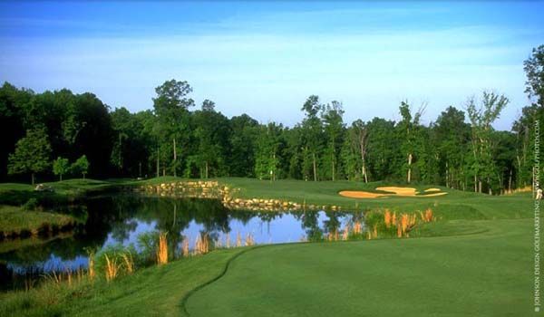 Golf course landscape with water hazard, trees, and green grass under a blue sky.
