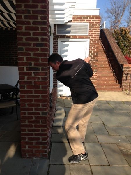 Man bends near brick building, looking downward. He wears a black jacket, tan pants, and dark shoes. Sunny outdoor setting.
