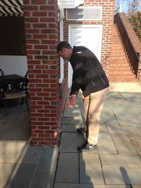 Man examining the edge of a brick building. He is wearing tan pants and a black jacket, standing on gray stone tiles.