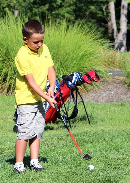 Boy in yellow shirt and shorts about to swing a golf club on a green lawn.