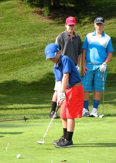 Boy in blue shirt and red shorts putting golf ball, two others watching on green.