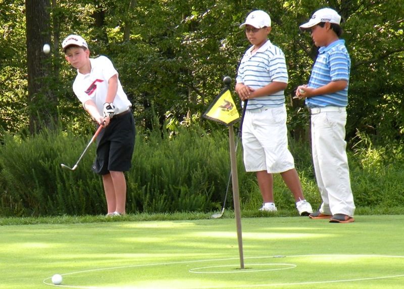 Boy swinging golf club on green, two others watch near flag.