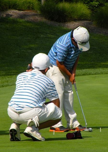 Two people putting on a golf course; one crouches, seemingly assisting the other with their shot.