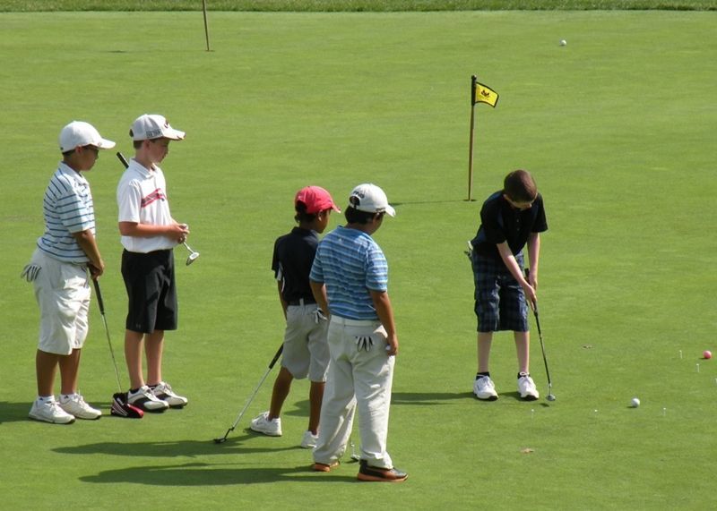 Five young golfers on a putting green; one puts, others watch with clubs, yellow flag in background.