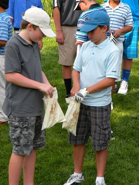 Two boys holding paper bags on a golf course, looking down. One wears a grey shirt, and the other a blue shirt.