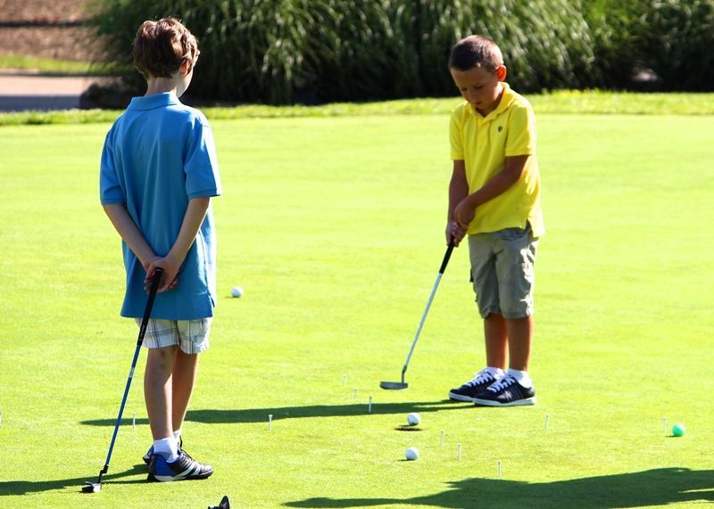 Two boys playing golf on a green course. One watches, one putts a ball.