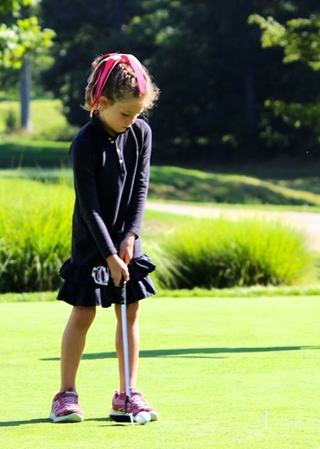 Girl putting a golf ball on a green, wearing a dark dress and pink sneakers.