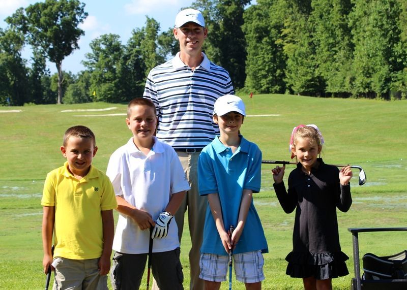 A man and four children stand on a golf course, holding clubs. Sunny day.
