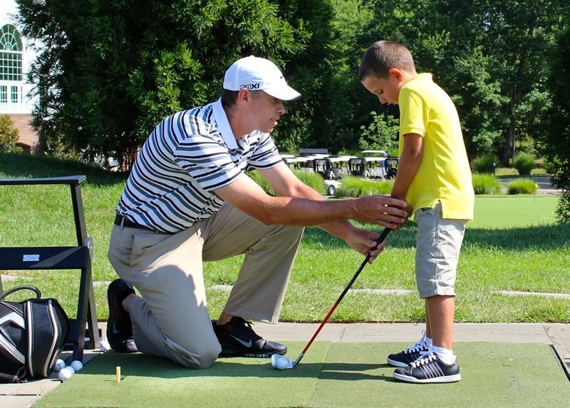 A man kneels, teaching a boy to golf. Outdoors, on a green mat. Man in striped shirt adjusts boy's grip.