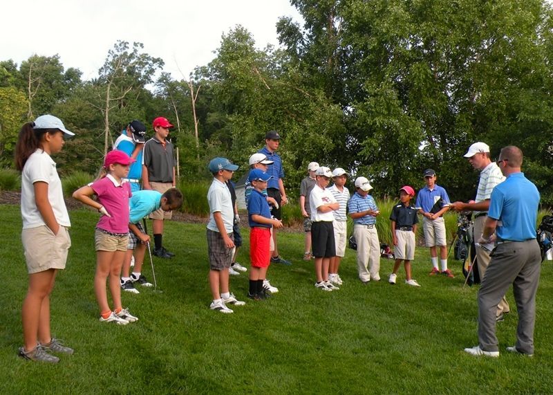 Group of children on a golf course listening to an instructor. Green grass, trees in the background.