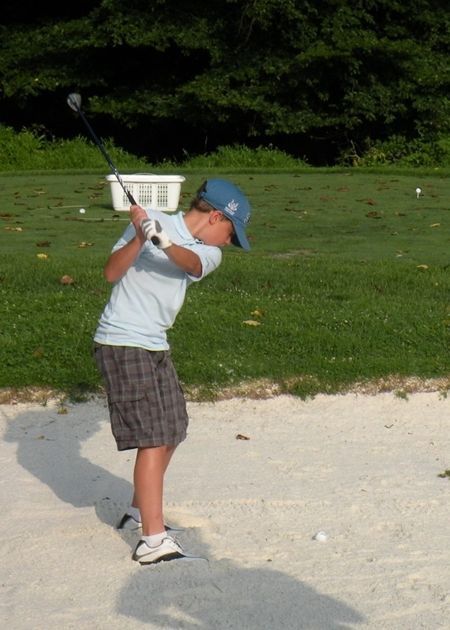 Boy in a blue hat swings a golf club in a sand trap on a golf course, with a bucket in the background.