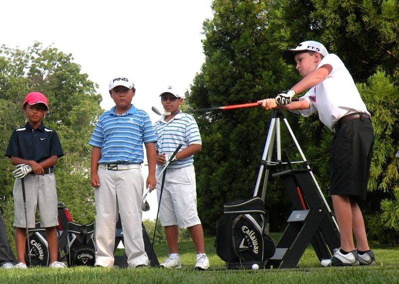 Four young golfers on a green field, one practicing swing with a training aid.