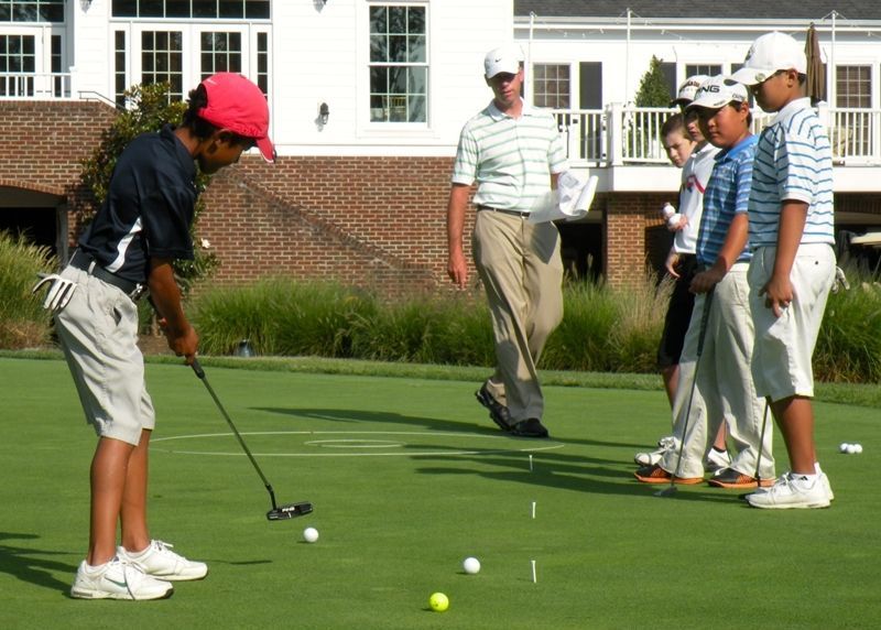 Boy in red hat putts golf ball on green with instructor and other children watching.