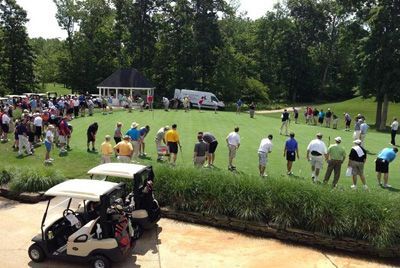 Golfers on putting green; two golf carts parked in foreground. Trees and small gazebo in background.