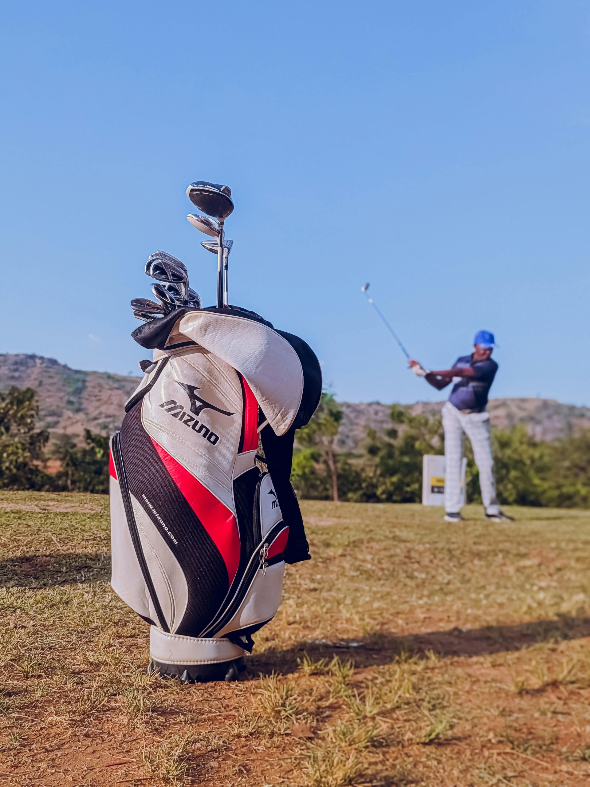 Golf bag in the foreground, golfer taking a swing in the background on a sunny day.