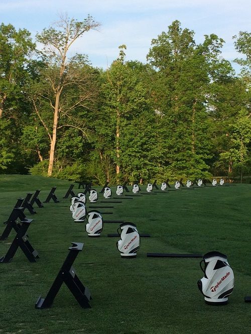 Golf driving range with white TaylorMade bags on green artificial turf. Trees in the background.