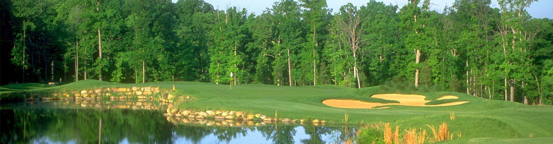 Golf course hole with a pond in the foreground and trees in the background. Green grass and sand trap.